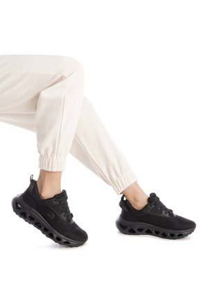 Zapatillas deportivas negras con una parte superior flexible y texturizada, una suela acolchada con agujeros de aire, y cordones para un ajuste seguro, combinadas con pantalones de chándal blancos.