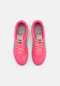 Klare rosa sneakers med grønn akcent, med en teksturert gummisåle, snøring, og mønstret innersåle. Puma-logo på tungen.