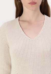 Pull en tricot beige avec un design en V, présentant un motif texturé et un poids moyen. Détails de couture près de l'épaule.