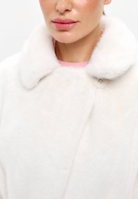 Vit fuskpälsjacka med bred krage, som har en rund knappstängning. Mjuk textur, enkel design och ren skärning.