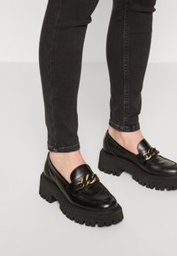 Mocassins noirs à talons épais avec détail de chaîne dorée, portés avec un jean skinny noir sur un sol gris clair.
