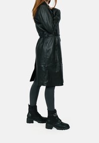 Gabardina negra de cuero con cintura ceñida con cinturón, llevada sobre leggings grises acanalados y botas negras gruesas con detalle de hebilla.