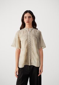Light beige blouse with short flutter sleeves, floral embroidery, narrow front placket, and decorative button details along the center.