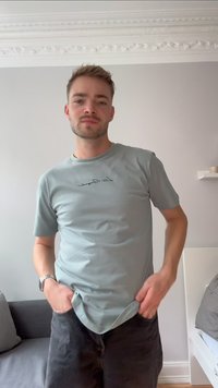 T-shirt di cotone verde chiaro con collo rotondo e maniche corte, caratterizzata da una scritta nera sul petto. Indossata con pantaloni corti scuri.