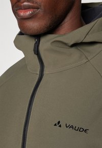 Olivegrüne Softshell-Jacke mit hohem Kragen, Reißverschluss vorne und schwarzem Logodetail. Sie verfügt über eine glatte Textur und eine taillierte Passform.