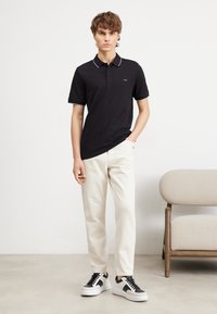Black polo shirt with white collar trim, paired with cream trousers and black-and-white sneakers. Simple design, casual fit.