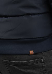 Tmavomodrá prešívaná bunda s textúrovaným povrchom, vybavená rebrovaným lemom a malou hnedou koženou značkou. Nosená cez čierne džínsové nohavice.