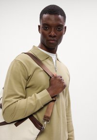 Hosszú ujjú, olíva zöld pulóver, cipzáros gallérral. A modellen világosbézs válltáska van, barna pántokkal és részletekkel. Sima textúra.