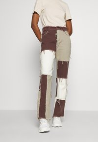 Lappade byxor med bruna, beige och vita tyger med fransade kanter. Hög midja-design. I kombination med vita sneakers.