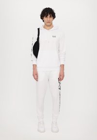Hvid hættetrøje med en lomme foran, snørebånd og logo på brystet. Parret med matchende hvide sweatpants med en lodret logo print.