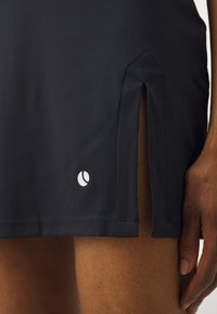 Μαύρη ραφτή αθλητική φούστα με πλαϊνές σχισμές και ένα μικρό λευκό λογότυπο στο κάτω μέρος εμπρός. Απαλή υφή με ορατές λεπτομέρειες ραφής.