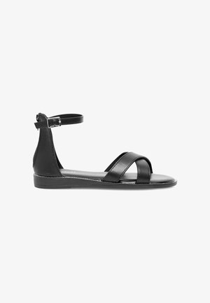 Schwarze flache Sandale mit Knöchelriemen, seitlicher Schnalle und zwei sich überkreuzenden Riemen im Zehenbereich vor weißem Hintergrund.