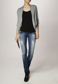 Grå öppen cardigan över en svart linne, i kombination med slitna blå denimjeans och svarta ankelstövlar; släta texturer och figurnära passform.