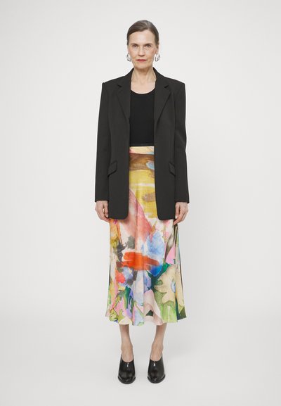 Blazer noir sur un haut noir ajusté, assorti à une jupe fluide à motifs floraux multicolores. Des chaussures à talons noirs complètent la tenue. Boucles d'oreilles en argent en forme de cerceau.