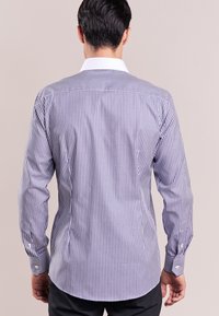 Uomo di spalle che indossa una camicia a maniche lunghe a righe verticali bianche e blu con colletto bianco, abbinata a pantaloni scuri.