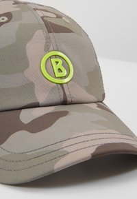 Casquette de baseball camouflage beige et marron avec une visière incurvée et un logo circulaire vert vif arborant la lettre « B » à l'avant.
