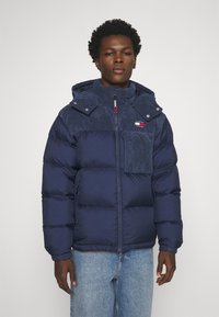 Giubbotto imbottito blu navy con parte superiore in corduroy, chiusura con zip, cappuccio e toppe con logo. Presenta sezioni imbottite e polsini elastici per garantire calore.