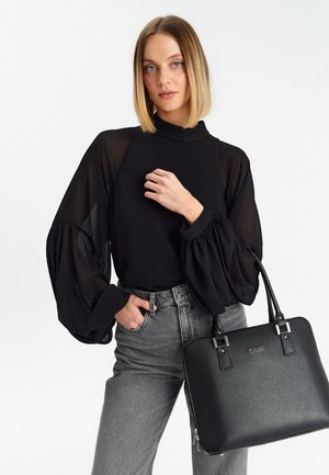 Top nero trasparente a maniche lunghe con maniche voluminose, indossato con jeans grigi a vita alta. Tiene in mano una borsa nera con texture e dettagli argento.