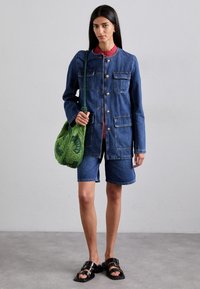 Denim jakna z zlatimi gumbi, štirimi sprednjimi žepi, v kombinaciji z denim kratkimi hlačami. Zeleni teksturiran nahrbtnik in črne sandale dopolnjujejo videz.