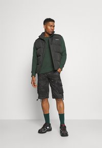 Gilet imbottito nero sopra un felpa verde, abbinato a pantaloni cargo camouflage neri. Calzature sono scarpe da ginnastica nere con accenti rossi.