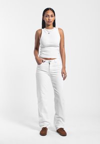 Top de tirantes blanco acanalado combinado con jeans blancos de pierna recta. La modelo lleva mocasines de ante marrón. Diseño simple, sin patrones ni acentos.