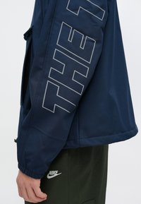 Tengerészkék könnyű dzseki nagy, szürke "THE" felirattal az ujjon, gumírozott mandzsettákkal és sima felülettel.