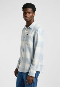 Camisa de cuadro azul claro con cuello abotonado, hecha de tela suave. Presenta un bolsillo en el pecho y detalles de botones en contraste.