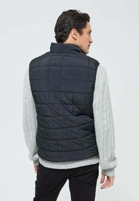 Gilet noir matelassé avec col, manches en tricot gris à motif torsadé et ourlet côtelé. Design léger et superposé pour une chaleur optimale.