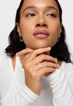Mujer con cabello largo y oscuro, lleva aretes de aro plateados y un anillo plateado con forma de rostro, apoyando su barbilla en sus manos entrelazadas.