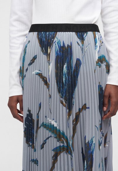 Gros plan sur une jupe plissée avec des motifs floraux abstraits bleus et bruns et une ceinture noire portée avec un haut blanc à manches longues.