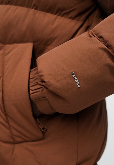 Brun pufferjakke med et elegant, broderet design. Har elastiske manchetter og et sølvfarvet "SANDRO" logo på ærmet. Tekstureret, let stof.