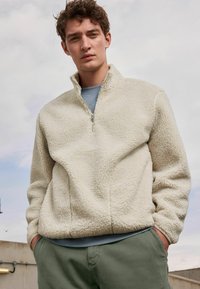 Pull en polaire crème avec un col zippé, texture douce, coupe décontractée, associé à un t-shirt bleu clair et un pantalon vert olive.