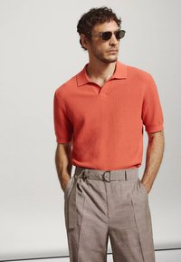 Homme portant des lunettes de soleil foncées, un polo corail texturé à manches courtes et un pantalon marron clair taille haute avec une ceinture, mains dans les poches.