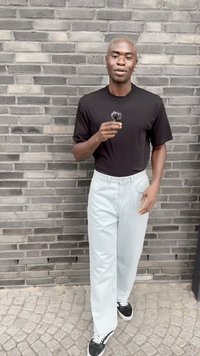 Mustusega lühikeste varrukatega särk, mis on kombineeritud helesiniste laia säärega pükste ja mustade spordijalatsitega valgete paeltega, seistes halli telliskiviseina vastu.