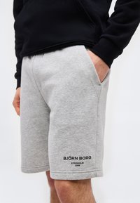 Άνδρας που φοράει ανοιχτά γκρι σορτς με το λογότυπο Björn Borg, με τα χέρια στις τσέπες και μαύρο μακρυμάνικο μπλουζάκι, μπροστά σε απλό φόντο.