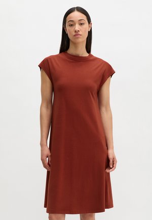 Kobieta z długimi ciemnymi włosami w bezrękawnikowej, rdzawo-kolorowej sukience do kolan stojąca na jednolitym białym tle.