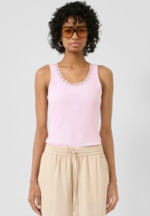 Femme aux cheveux bouclés portant de grandes lunettes teintées ambre, un débardeur rose bordé de dentelle, et un pantalon beige texturé avec un cordon de serrage.