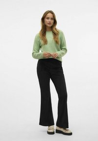 Maglione verde chiaro in morbido tessuto, abbinato a pantaloni neri a zampa d'elefante e scarpe beige con dettagli frangiati, in piedi davanti a uno sfondo semplice.