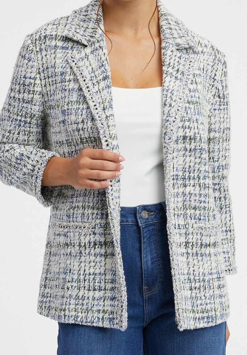 Femme portant un blazer en tweed texturé bleu et blanc sur un top blanc et un jean bleu, tenant le revers du blazer de la main gauche.