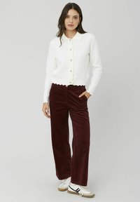 Cardigan blanc côtelé avec un ourlet festonné et de gros boutons, associé à un pantalon large en velours côtelé bordeaux. Le modèle porte des baskets blanches.