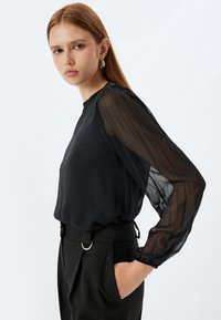Blusa nera con maniche lunghe trasparenti e plissettate; corpo liscio; abbinata a pantaloni a righe sottili; indossata con un sottile anello in accentuazione in vita.