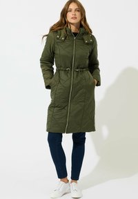 Mujer de pie vistiendo un abrigo largo acolchado verde olivo con cierre delantero, jeans oscuros y zapatillas blancas contra un fondo blanco.