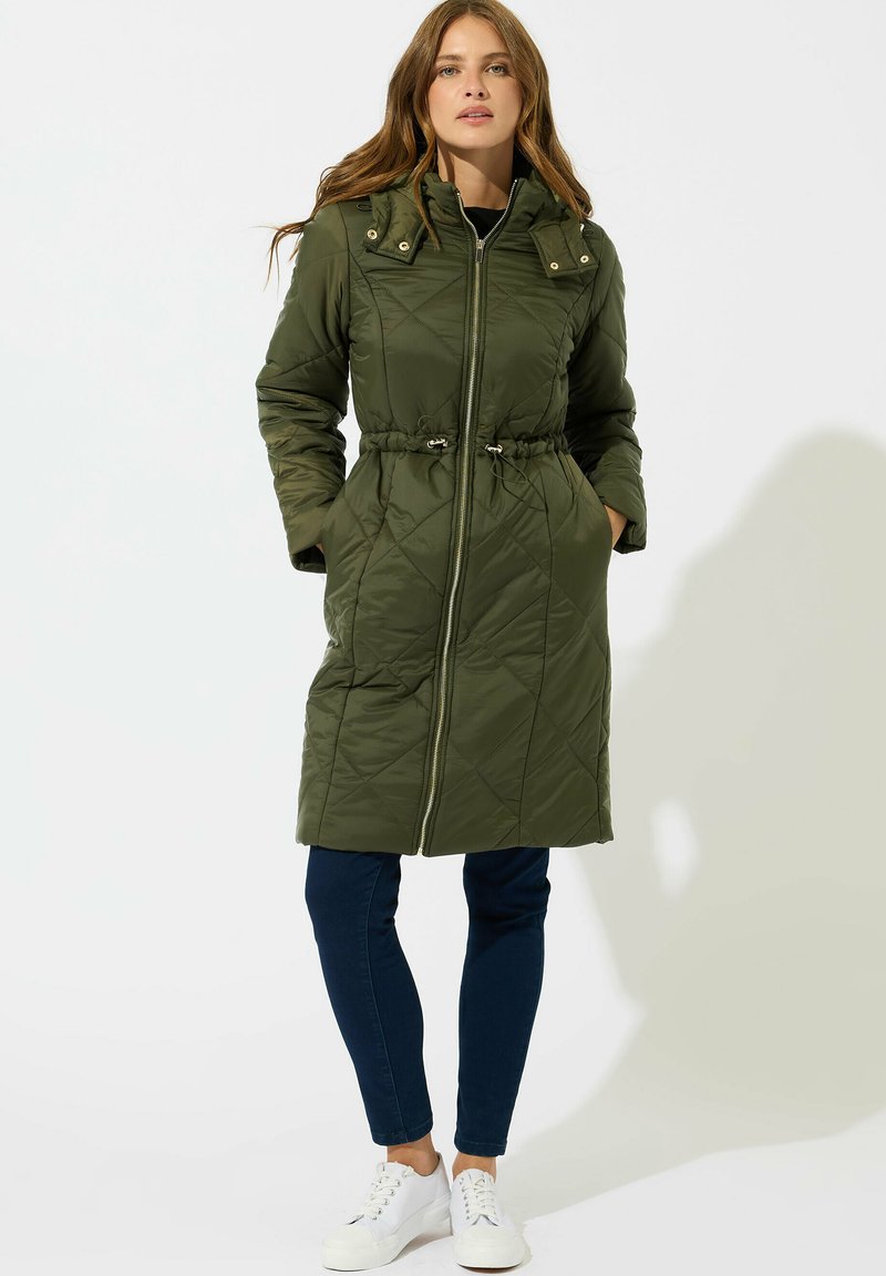 Mujer de pie vistiendo un abrigo largo acolchado verde olivo con cierre delantero, jeans oscuros y zapatillas blancas contra un fondo blanco.