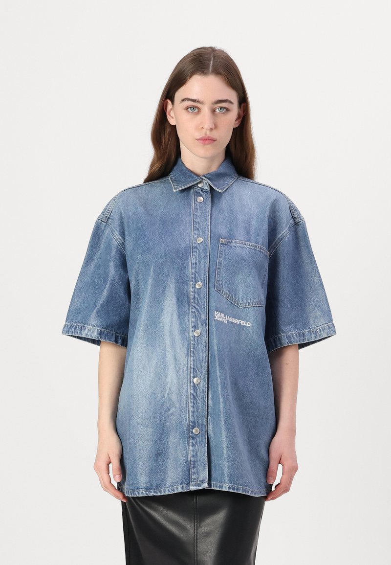 Μπλε τζιν oversized πουκάμισο με κοντά μανίκια, κουμπιά μπροστά και τσέπη στο στήθος. Διαθέτει λεπτομέρεια λογότυπου στο μπροστά μέρος.