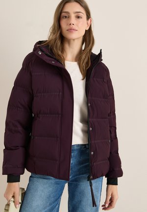 Veste en duvet bordeaux avec capuche, design matelassé, fermeture éclair, et poignets en accent noir, associée à un pull léger et un jean bleu.