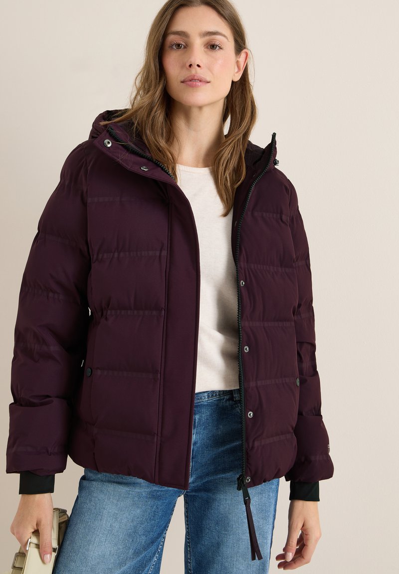 Giubbotto in piuma color marsala con cappuccio, design trapuntato, chiusura con zip e polsini neri, abbinato a un maglione leggero e jeans blu.