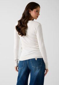 Weißer Pullover mit langen Ärmeln, einem gerippten Ausschnitt und Bündchen, kombiniert mit blauen Denim-Jeans mit einem dreieckigen Taschendetail.