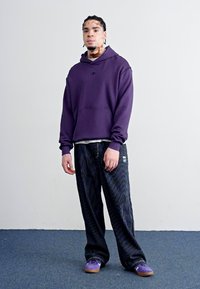 Lilla hettegenser med frontlomme, kombinert med brede bukser med svarte striper og lilla sko. Stoffet ser mykt ut med en avslappet passform.
