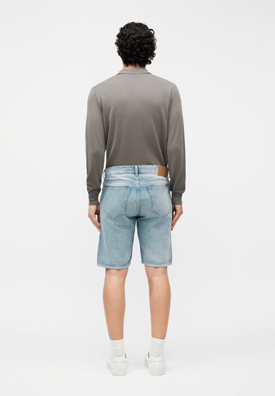 Άνδρας με σγουρά μαλλιά, φοράει γκρι μακρυμάνικο πουκάμισο, ανοιχτόχρωμα τζιν σορτς, λευκές κάλτσες και λευκά αθλητικά παπούτσια, και στέκεται με την πλάτη προς το μέρος μας.