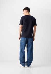 Tricou bleumarin cu mânecă scurtă purtat cu blugi albaștri cu talie înaltă și croială lejeră. Modelul poartă adidași albi, văzut din spate.